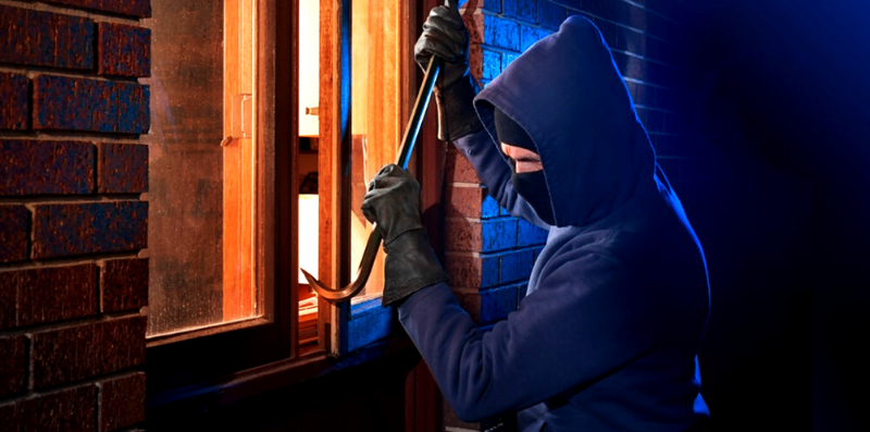 Burglar in black hoodie and a ski mask breaking into a home by a window with a crowbar